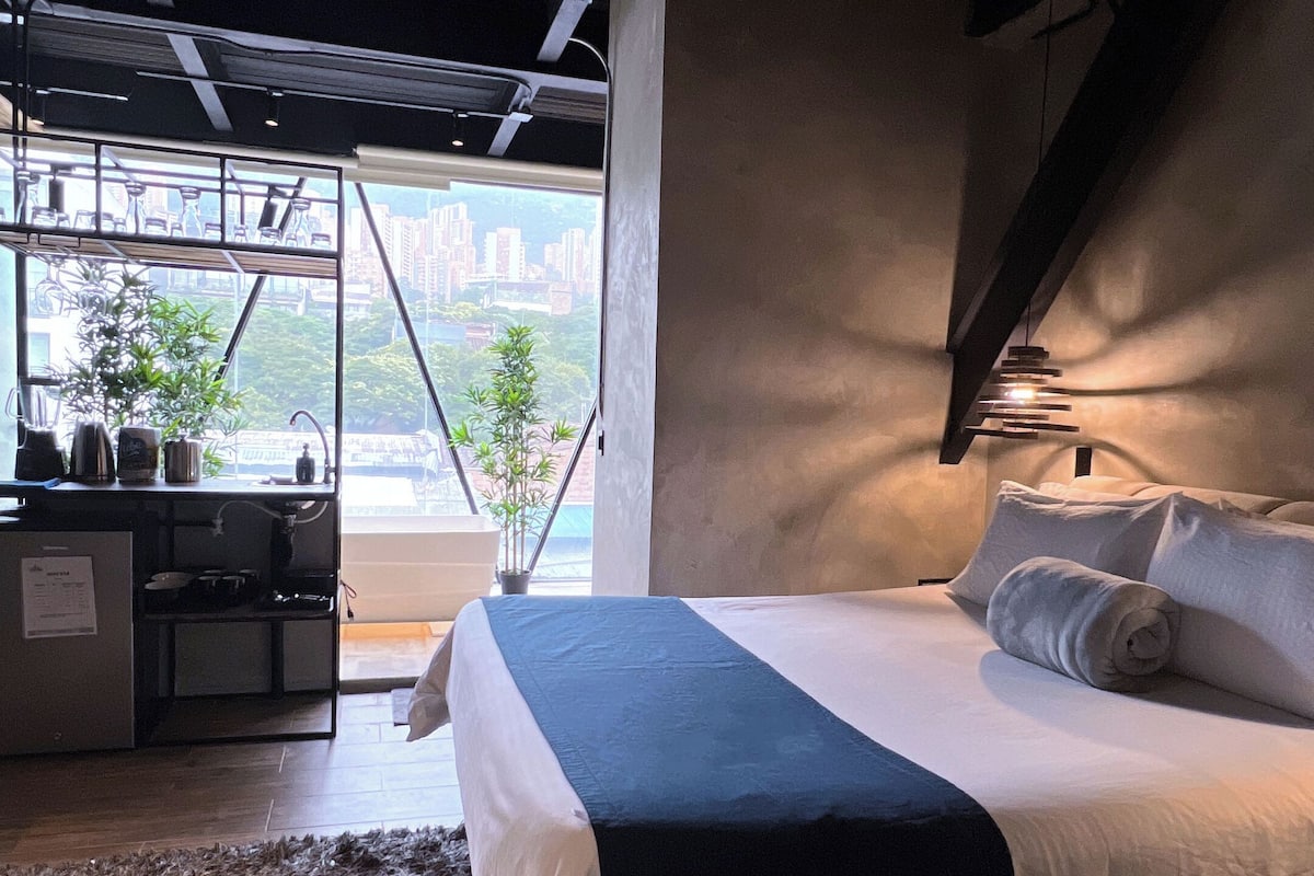 Ein modernes Schlafzimmer mit einem großen Bett, einem Schreibtisch und Blick auf die Stadt durch ein großes Fenster.
