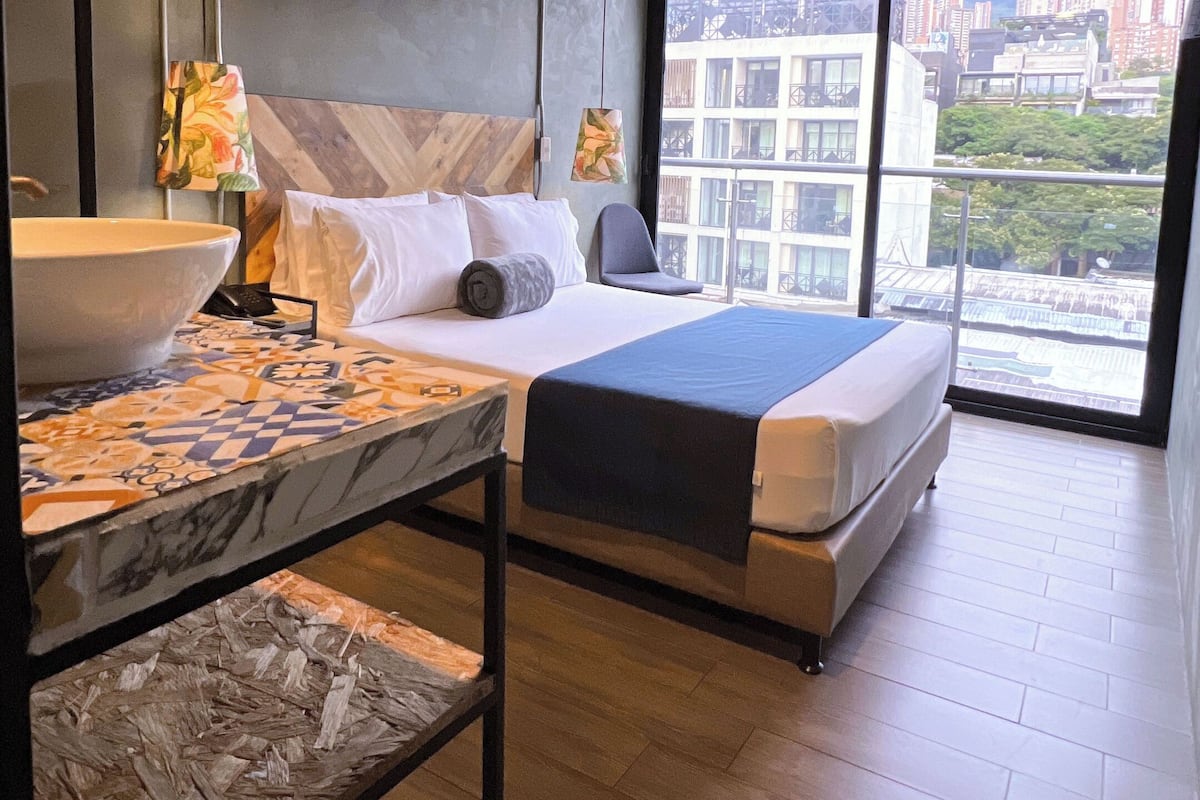 Ein modernes Hotelzimmer mit einem großen Bett, einem Tisch mit Marmorplatte und Blick auf die Stadt durch große Fenster.