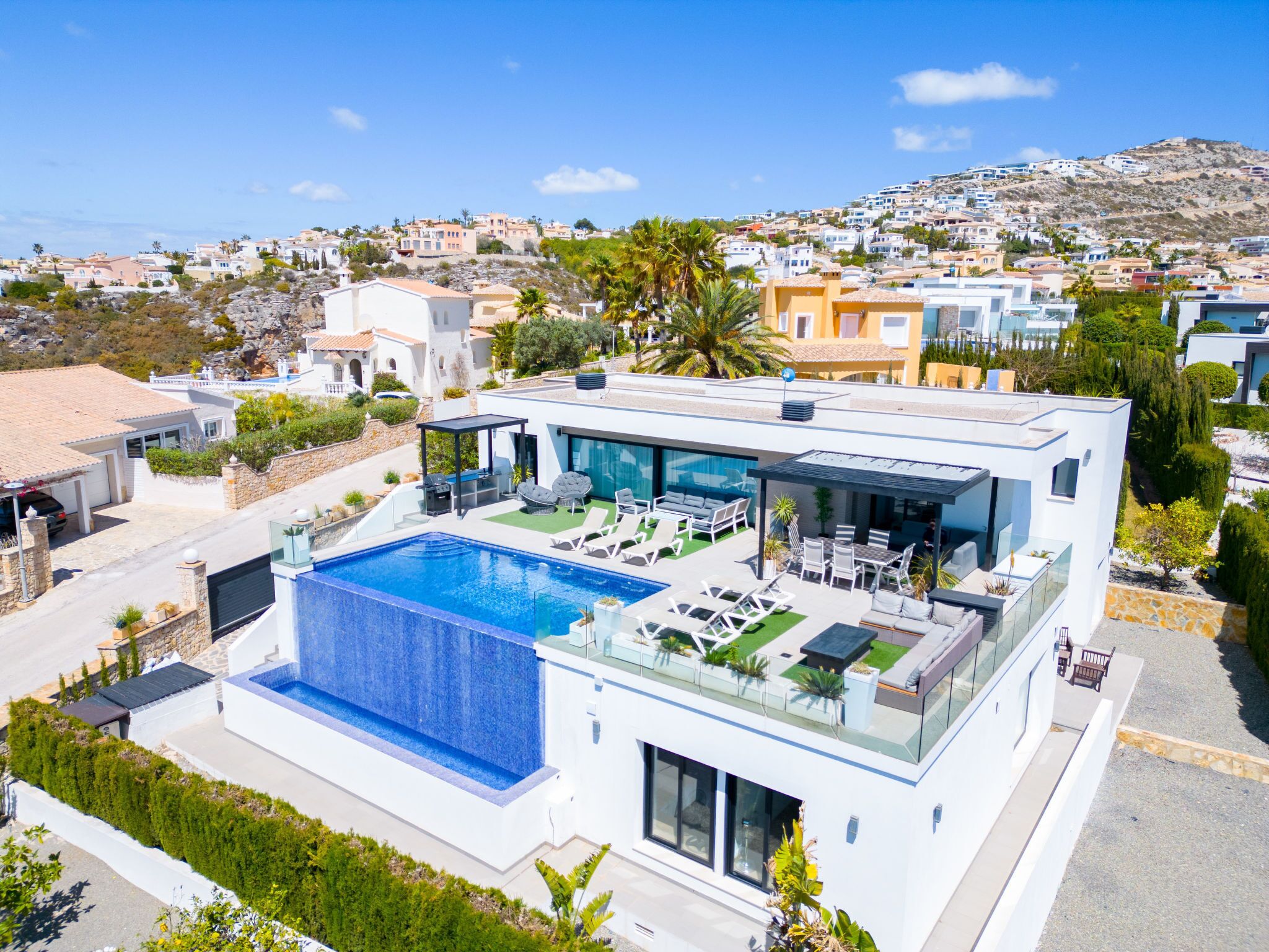 A modern villa with a large swimming pool, a covered outdoor seating area, and a view of the surrounding hillside neighborhood.