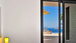A hotel room with a bed, a nightstand with a lamp, and a view of a beach with a chair and umbrella. - Vacation home Megin by Interhome (Moraira)