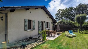 A house with a sloped roof, a small porch with a table and chairs, and a fenced yard with a bench and a hammock. - Gîte du Héron Gris (BOULIGNEUX)