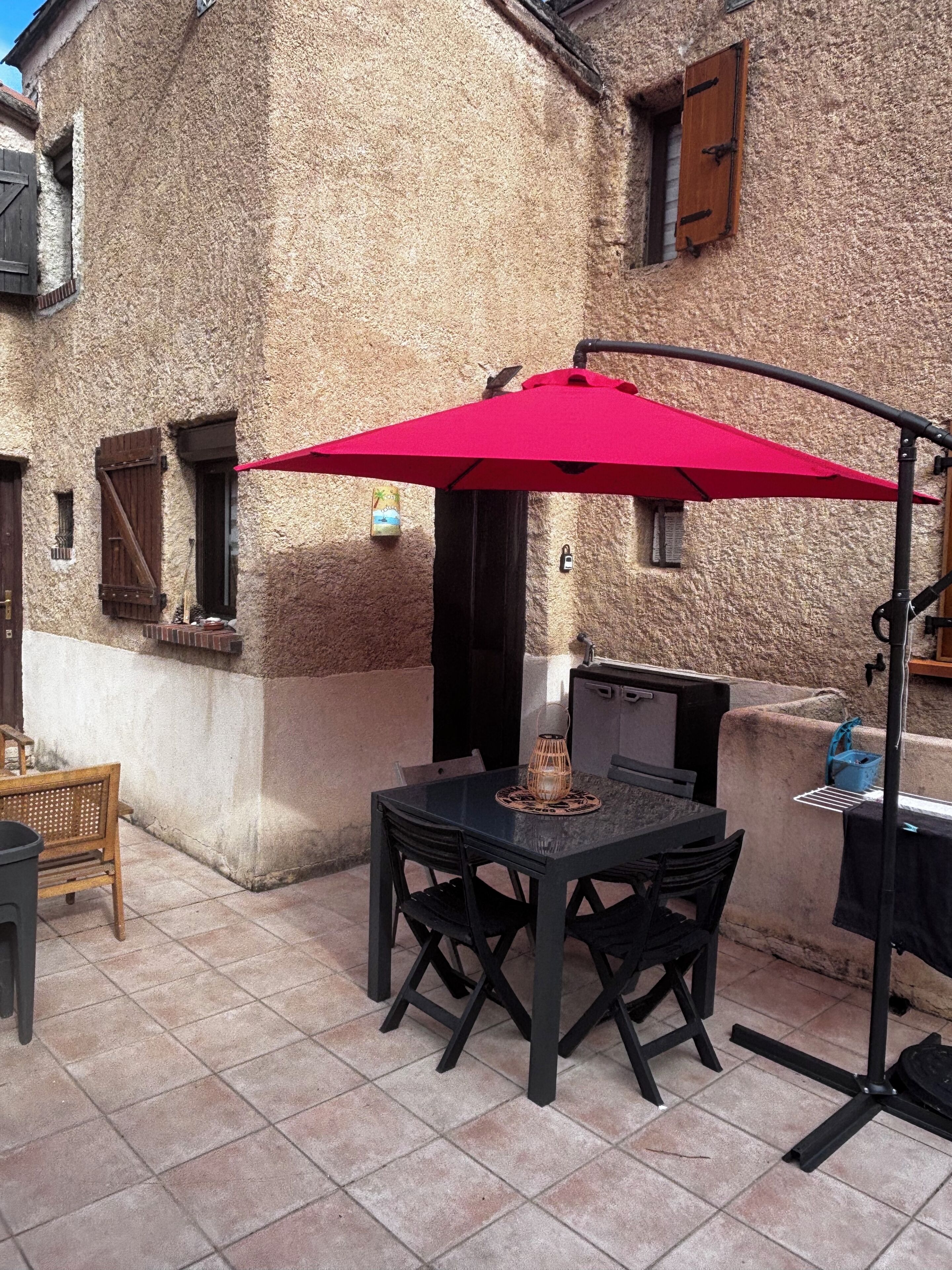 Une terrasse extérieure avec une table et des chaises, un parasol rouge et un bâtiment en pierre en arrière-plan.