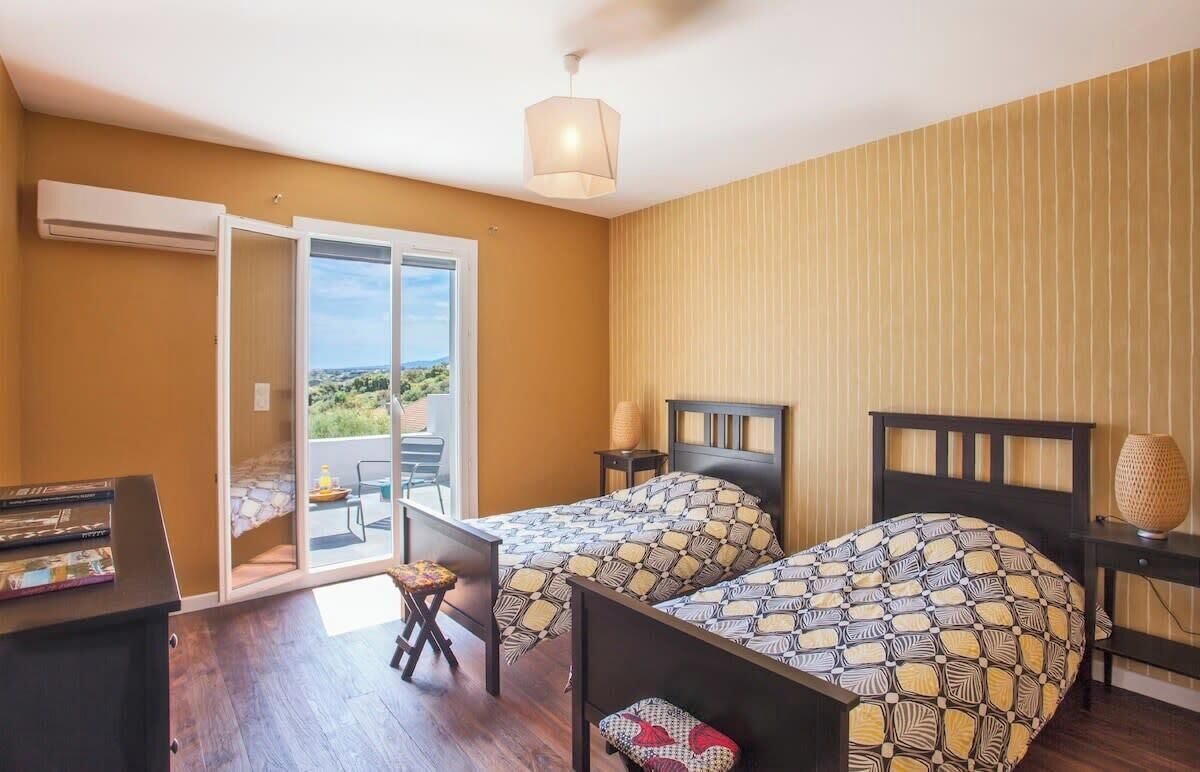 A room with two beds, a desk, and a chair. There is a window with a view of greenery and a balcony.