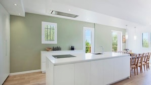 A modern kitchen with a large island, white cabinetry, and a view of the outdoors through a window. - Villa Le Corps de Garde (Furiani)