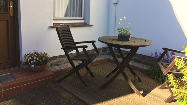 Eine Holzdar Terrasse mit Tisch und Stühlen, einem Blumentopf und einem Fenster mit Vorhang.