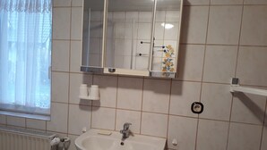 Ein Badezimmer mit einem weißen Waschbecken, einem Spiegel mit floralem Dekor und einem Fenster mit leichtem Vorhang.