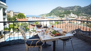 Une terrasse avec une table à manger dressée pour un repas, offrant une vue sur une ville côtière et les montagnes.