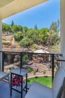 Un balcón con mobiliario de mimbre, una mesa de vidrio con flores y vista a un jardín ajardinado con árboles y una piscina.