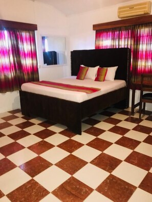 A bed with a dark headboard and multiple pillows, a checkered floor, and a window with curtains. - The Ivy Hotel Santa Maria (Accra)