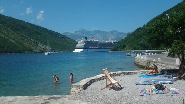 Een cruiseschip ligt aangemeerd in een haven met een kiezelstrand en enkele mensen in het water.
