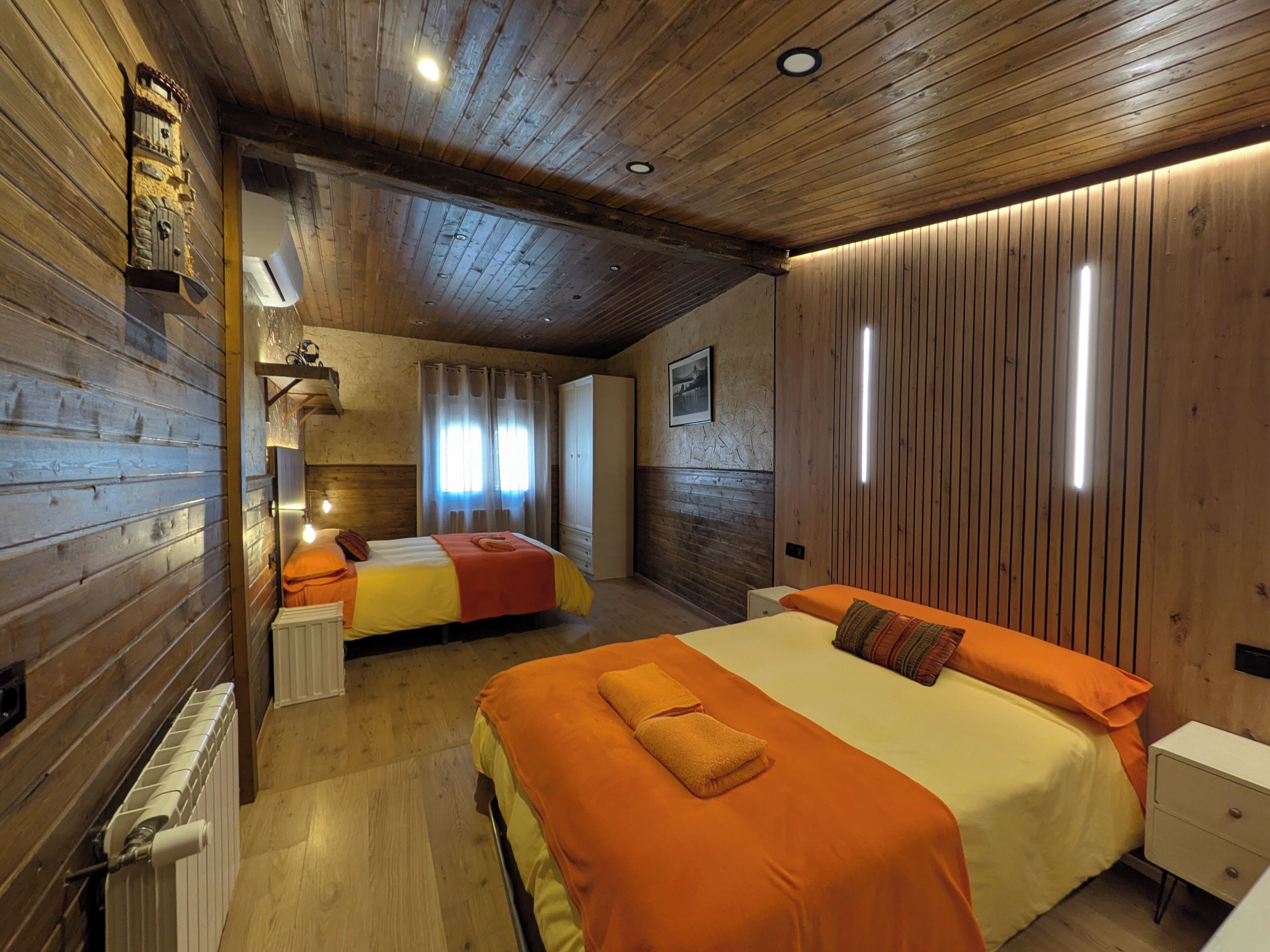 Una habitación con paneles de madera, dos camas con ropa de cama naranja, un radiador y un armario blanco.