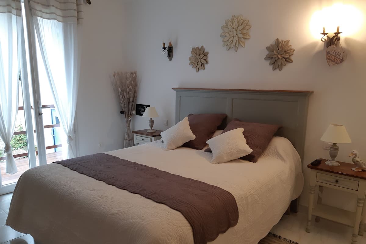 Un lit bien fait, recouvert d’un plaid marron, une table de chevet avec une lampe et des décorations florales accrochées au mur.