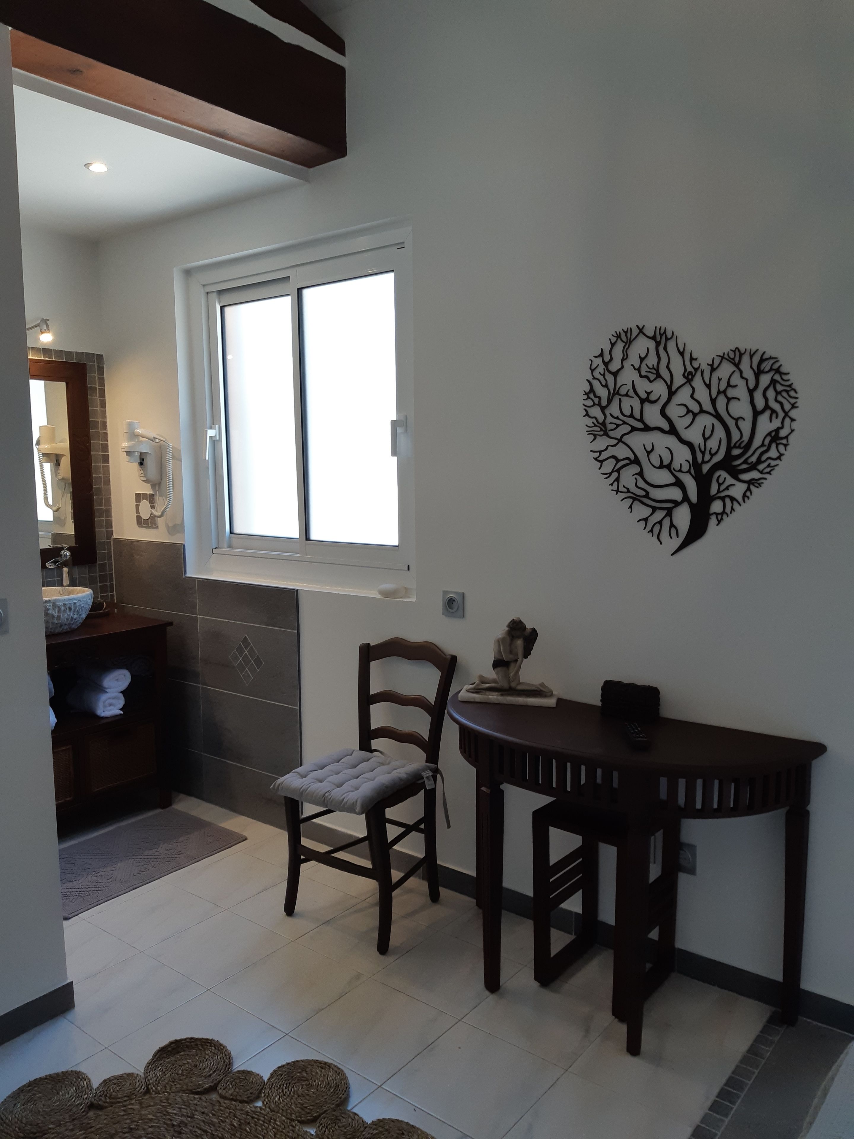 A room with a dining table, a chair, a wall-mounted artwork, a window, and a bathroom visible in the background.