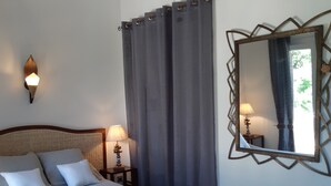A neatly made bed with pillows, a bedside lamp, a wall-mounted mirror, and a woven rug on the floor. - Chambres d'hotes - Villa la Licorne (Belcodene)