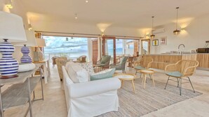 Un vaste espace de vie doté d’une grande fenêtre donnant sur la mer, aménagé avec des canapés blancs et des fauteuils en osier, une table à manger en bois et un coin cuisine avec un bar.