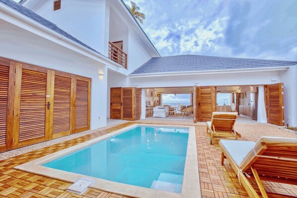 A pool area with wooden shutters, lounge chairs, and a view of the ocean.