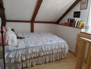 Een slaapkamer met een schuine kap, een bed met een sprei, een houten stoel, een plank met boeken en een aan de muur hangend schilderij.