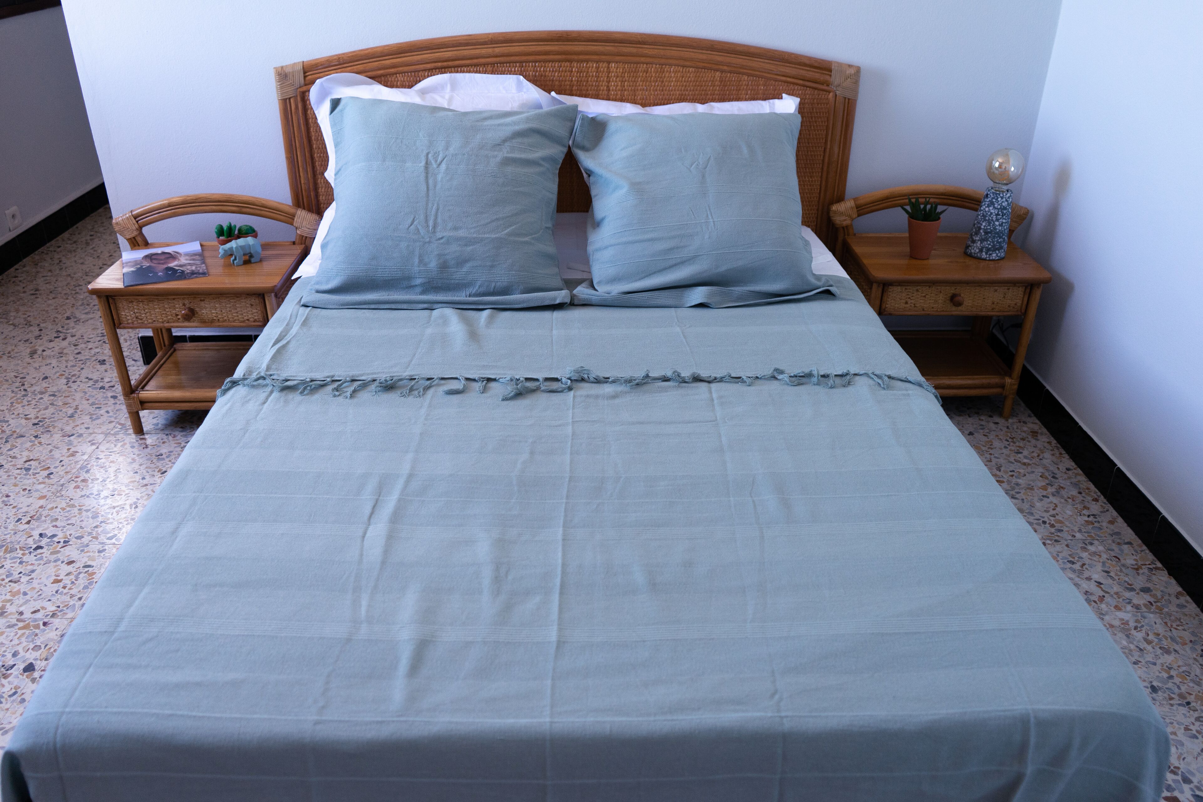 Een net opgemaakt bed met een lichtblauwe deken en kussens, aan weerszijden omringd door houten nachtkastjes met kleine potplanten en decoratieve items.
