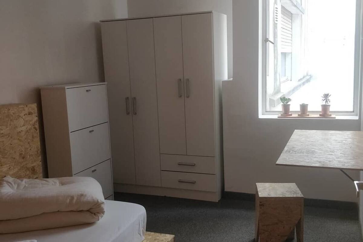 A bedroom with a bed, a wardrobe, a small table, and a window with a view of buildings.
