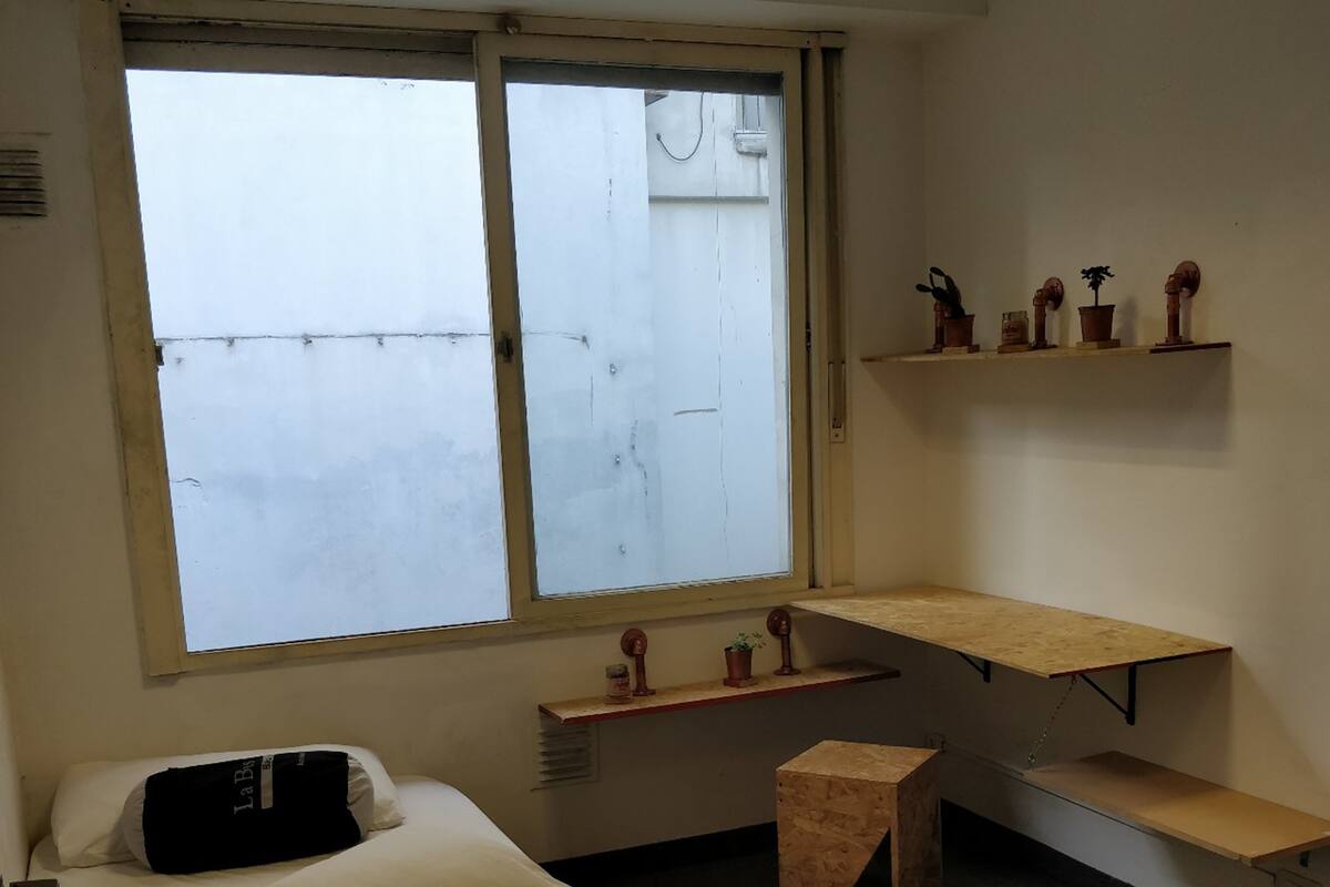 A small, simple room with a bed, a window, a shelf with decorative items, and a small stool.