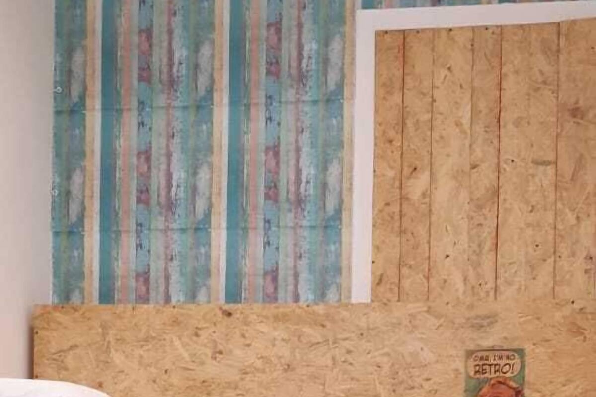 A room with a wooden bed frame, a small wooden shelf with a retro poster, and a patterned curtain.