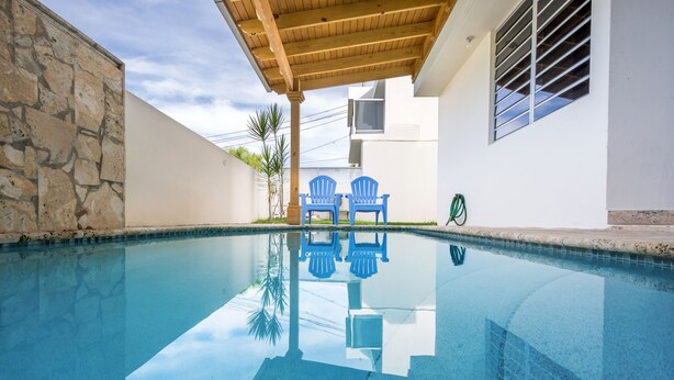 A swimming pool with clear blue water, two blue chairs, a stone wall, and a building with a white exterior and a wooden pergola. - Private pool, Oceanview, Centrally located! (Puerto)