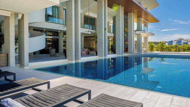 Area piscina moderna con sedie a sdraio, piscina cristallina e un edificio contemporaneo con ampie finestre e colonne.