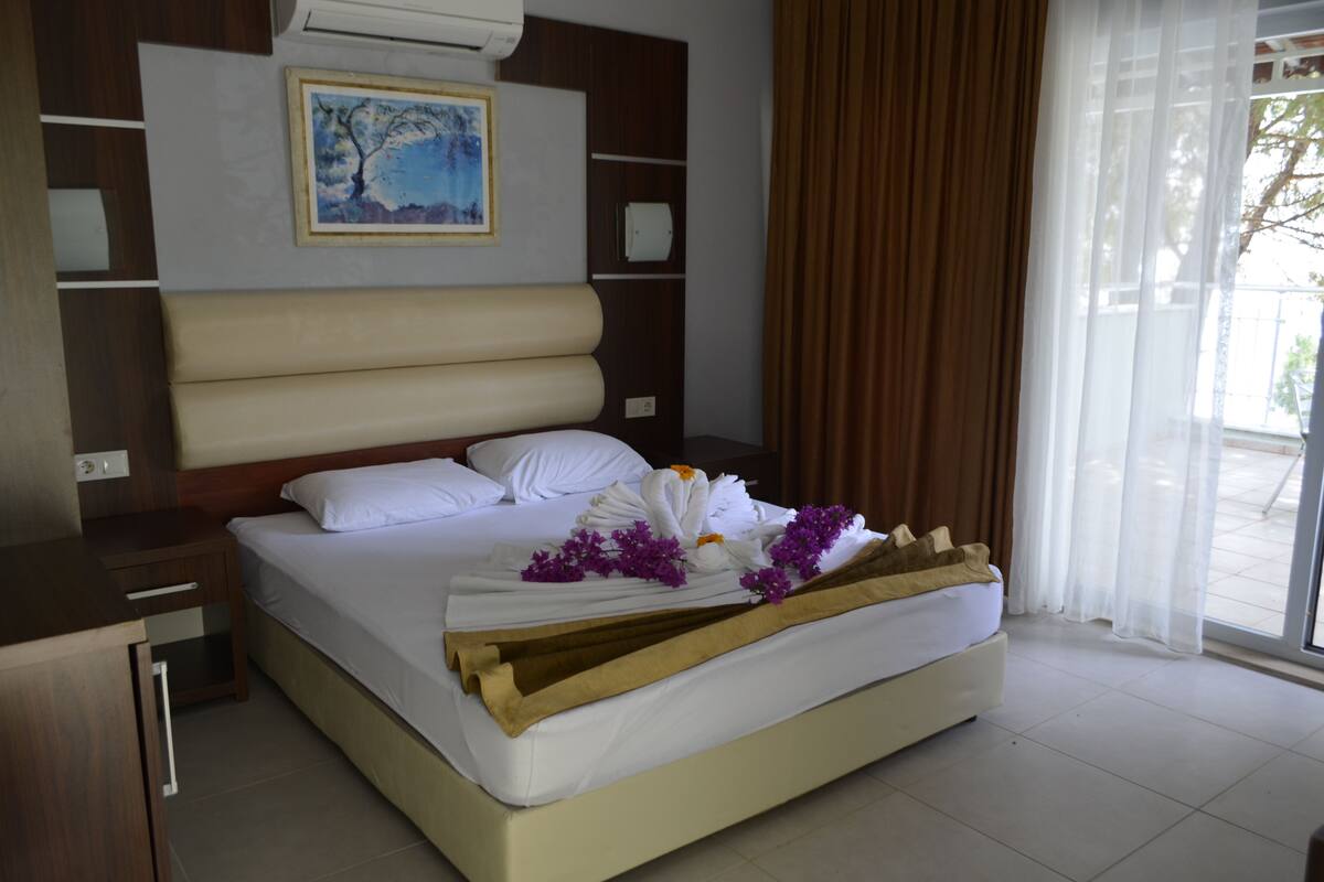 A hotel room with a large bed, a nightstand, a wall-mounted air conditioner, a painting, and a view through the window.