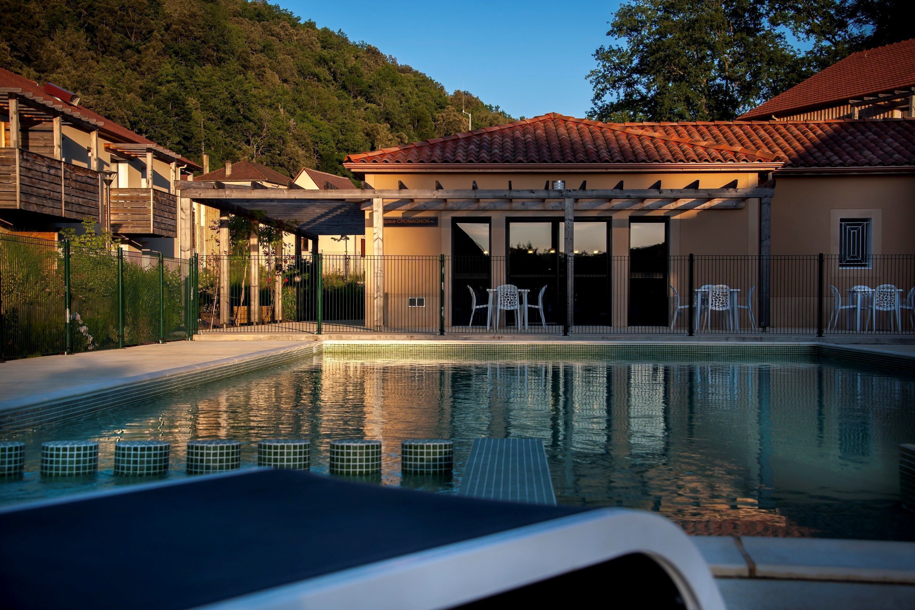 A swimming pool with tiled edges, a covered seating area with chairs, and a building with a tiled roof.