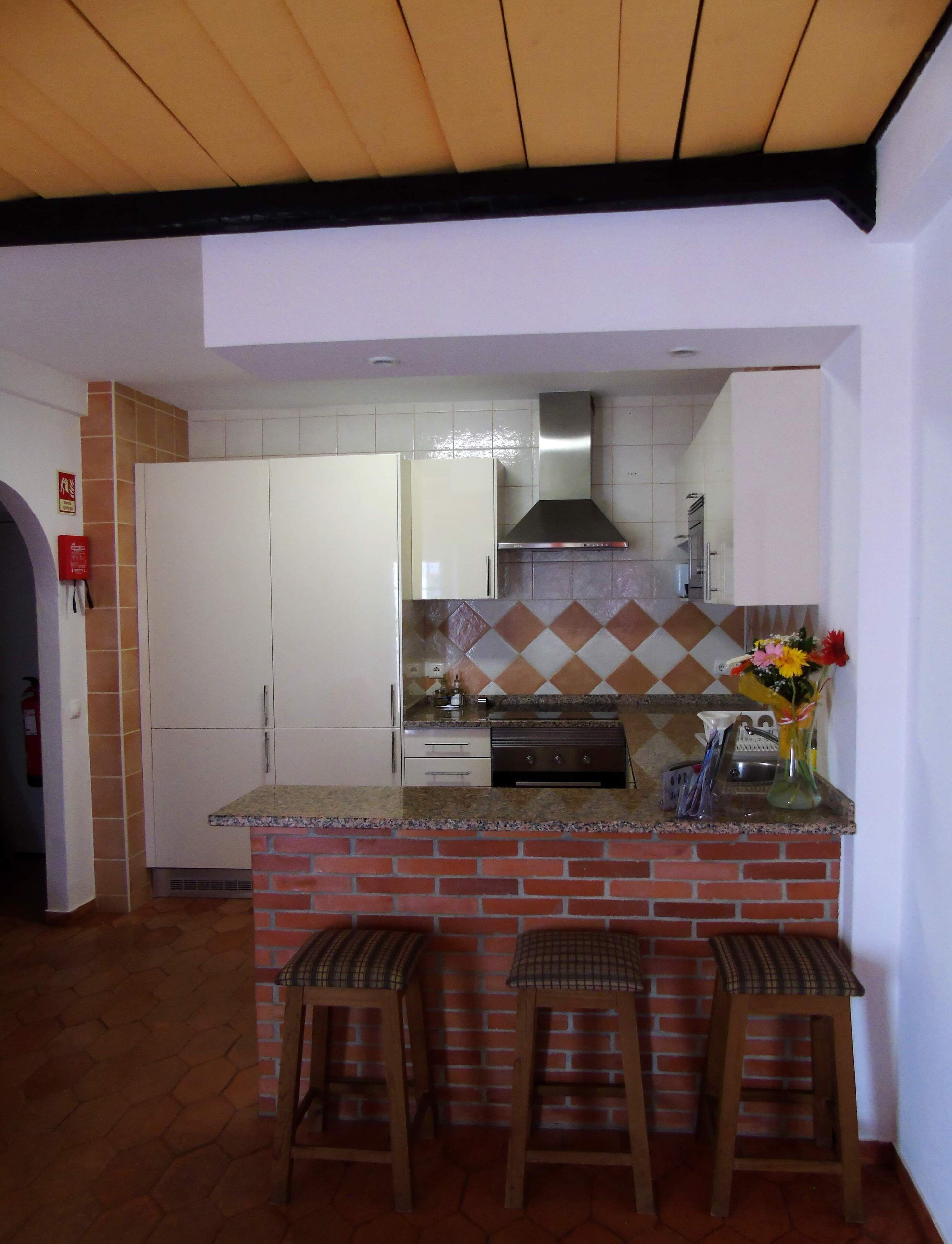 A kitchen with a brick island, white cabinets, a stainless steel range hood, and a tiled backsplash. There are three wooden stools at the island.