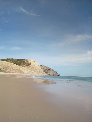 Une plage avec un rivage sablonneux, des vagues douces et une falaise rocheuse en arrière-plan.