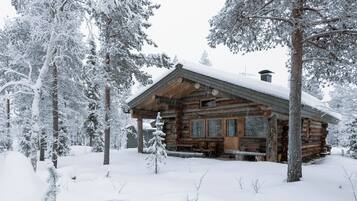 Lumihuntuun peittynyt metsä ympäröi hirsihökkelöä.