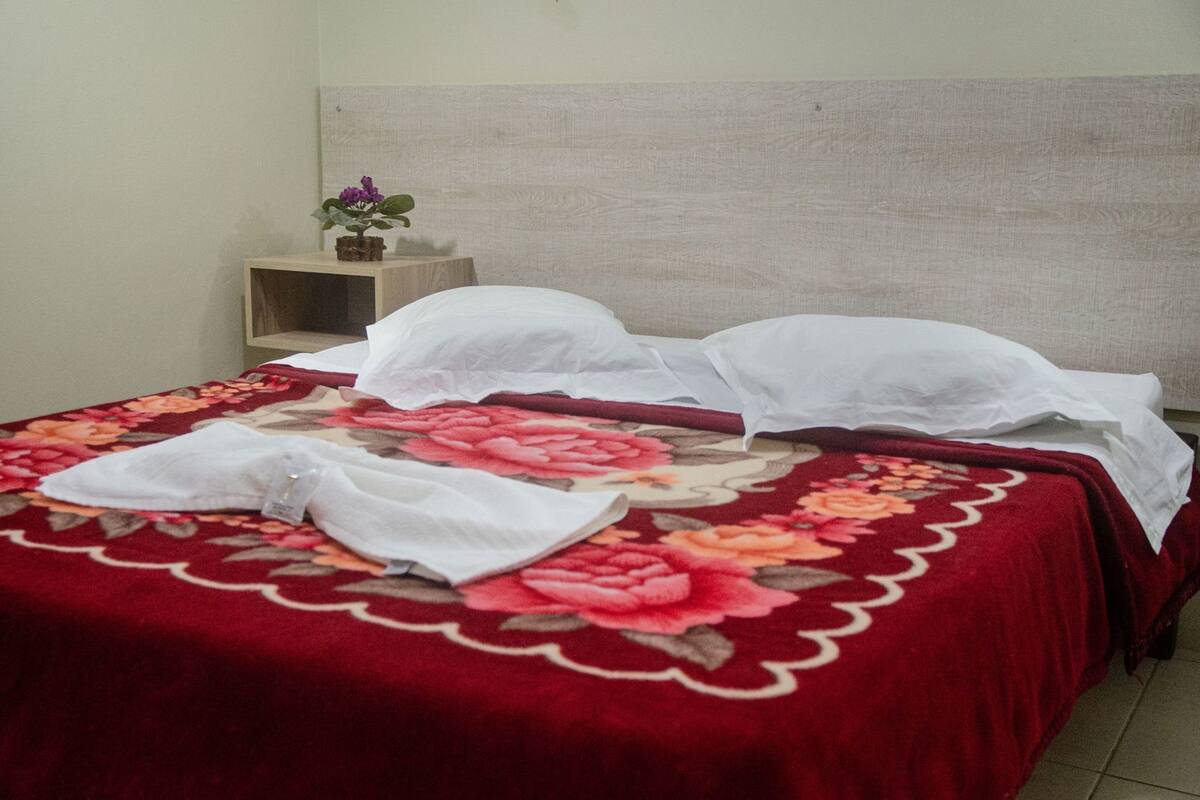 Un lit recouvert d’une couvre-lit à motifs floraux, une petite table de chevet en bois avec un pot de plante et une tête de lit sobre.