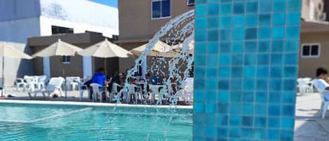 A swimming pool with clear water, surrounded by lounge chairs and umbrellas, with buildings in the background.