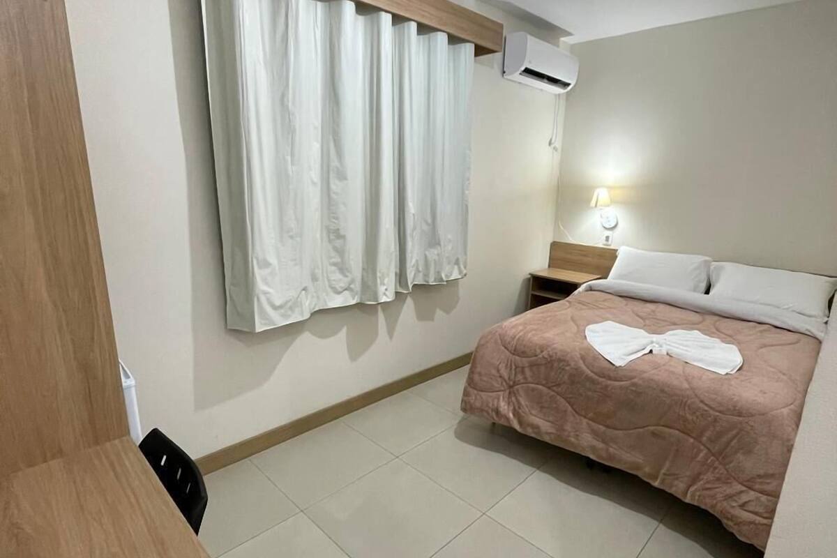 Une chambre d’hôtel comprenant un lit, un bureau, une chaise, une fenêtre avec des rideaux, un appareil de climatisation et une table de chevet avec une lampe.