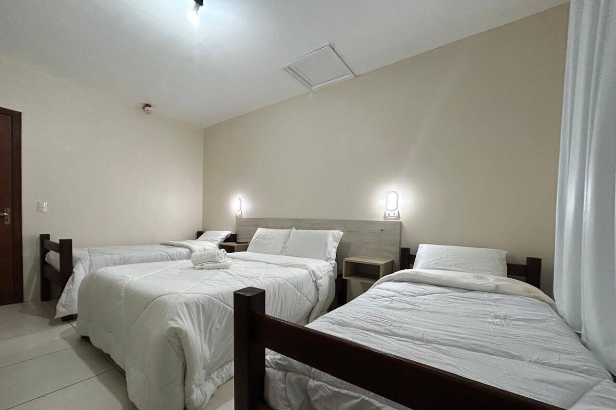 Une chambre d’hôtel avec trois lits simples, une tête de lit en bois, des tables de chevet et un rideau blanc.