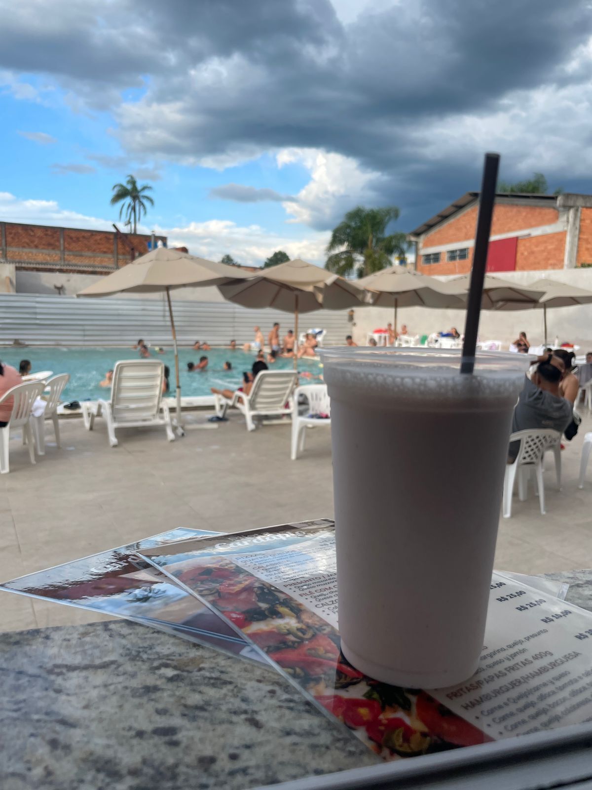 Une boisson servie dans un verre avec une paille, un menu et un espace piscine avec des personnes et des parasols.