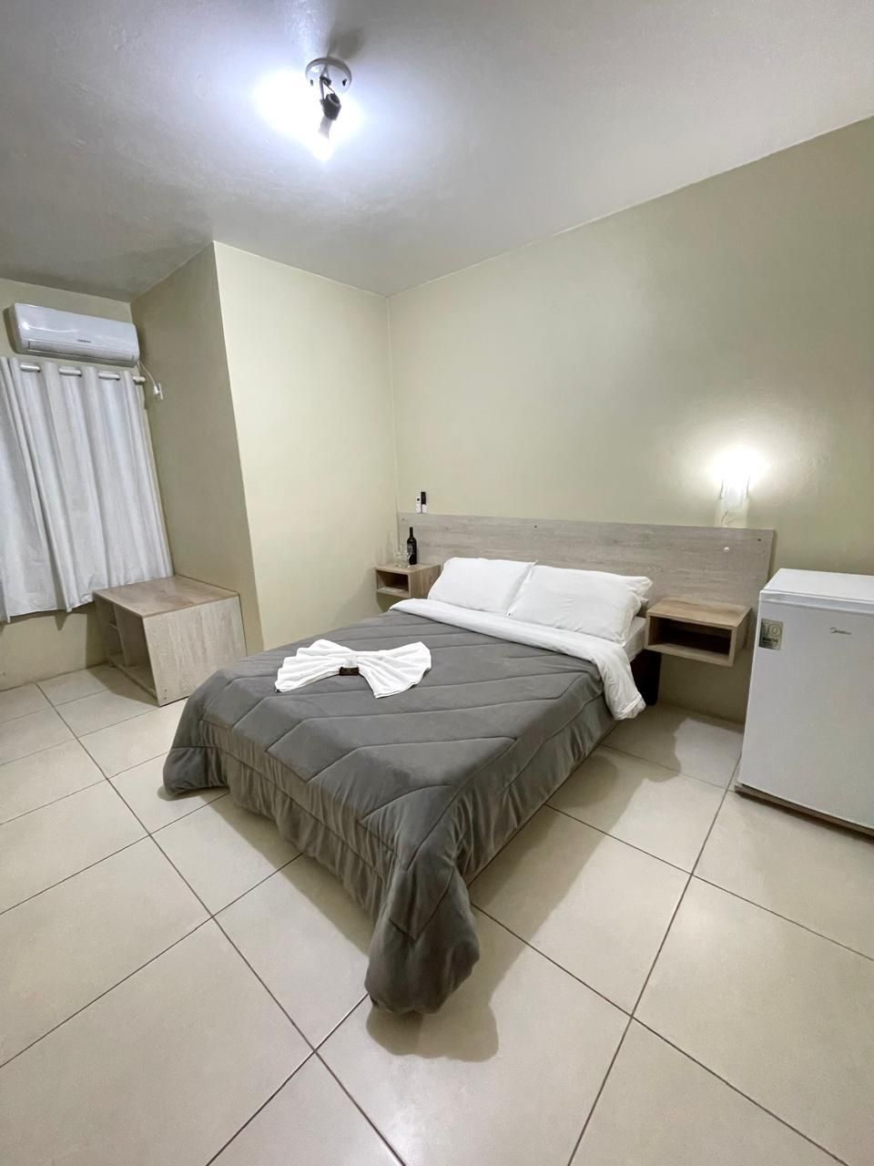 A bedroom with a bed, a nightstand, a wall-mounted heater, and a small refrigerator.