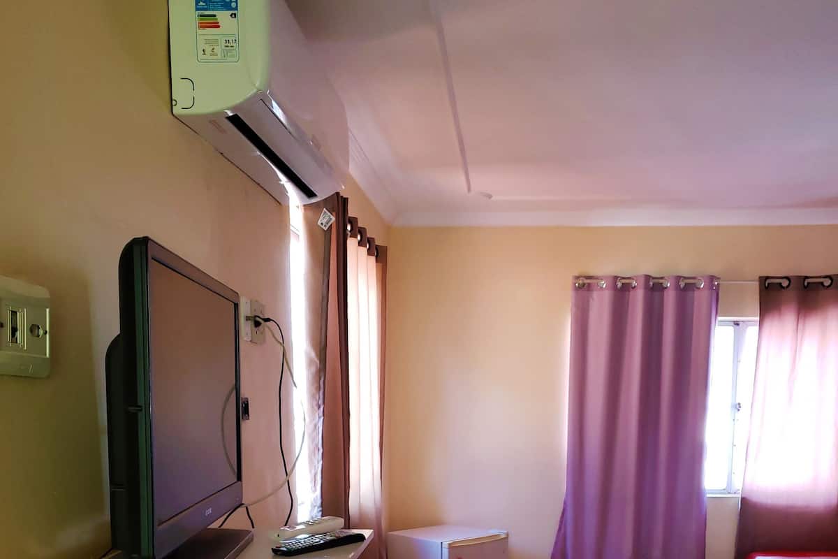 Ein Zimmer mit einem Bett, einem Schreibtisch mit Computer, einer kleinen Klimaanlage und einem Fenster mit Vorhängen.