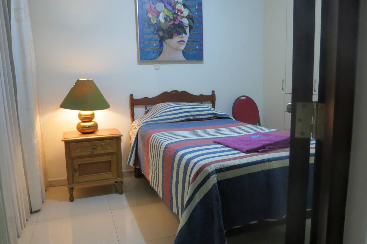 A bedroom with a bed, a nightstand with a lamp, a painting on the wall, and a door.