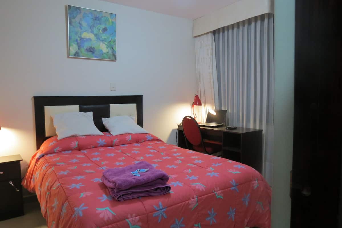 A hotel room with a bed, a desk, a chair, a lamp, and a painting on the wall.