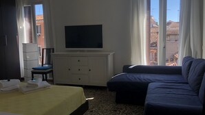 A hotel room with a bed, a sofa, a TV on a cabinet, and a window with a view of buildings. - Al Teatro Italia (Venice)