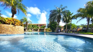 A clear swimming pool surrounded by palm trees and a stone wall, with a building in the background. - Eagle beach walking distance Townhouse (Oranjestad-West)