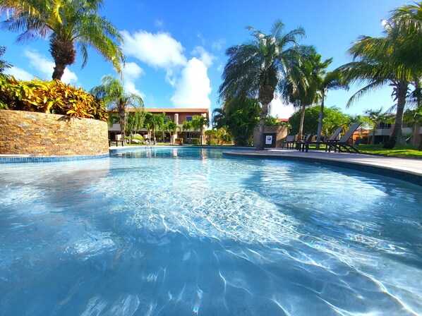 A clear swimming pool surrounded by palm trees and a stone wall, with a building in the background.