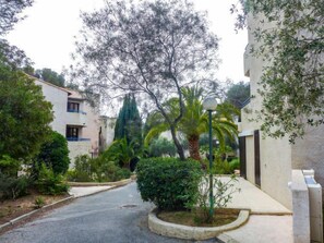 A narrow street flanked by buildings with balconies, surrounded by lush greenery and palm trees.