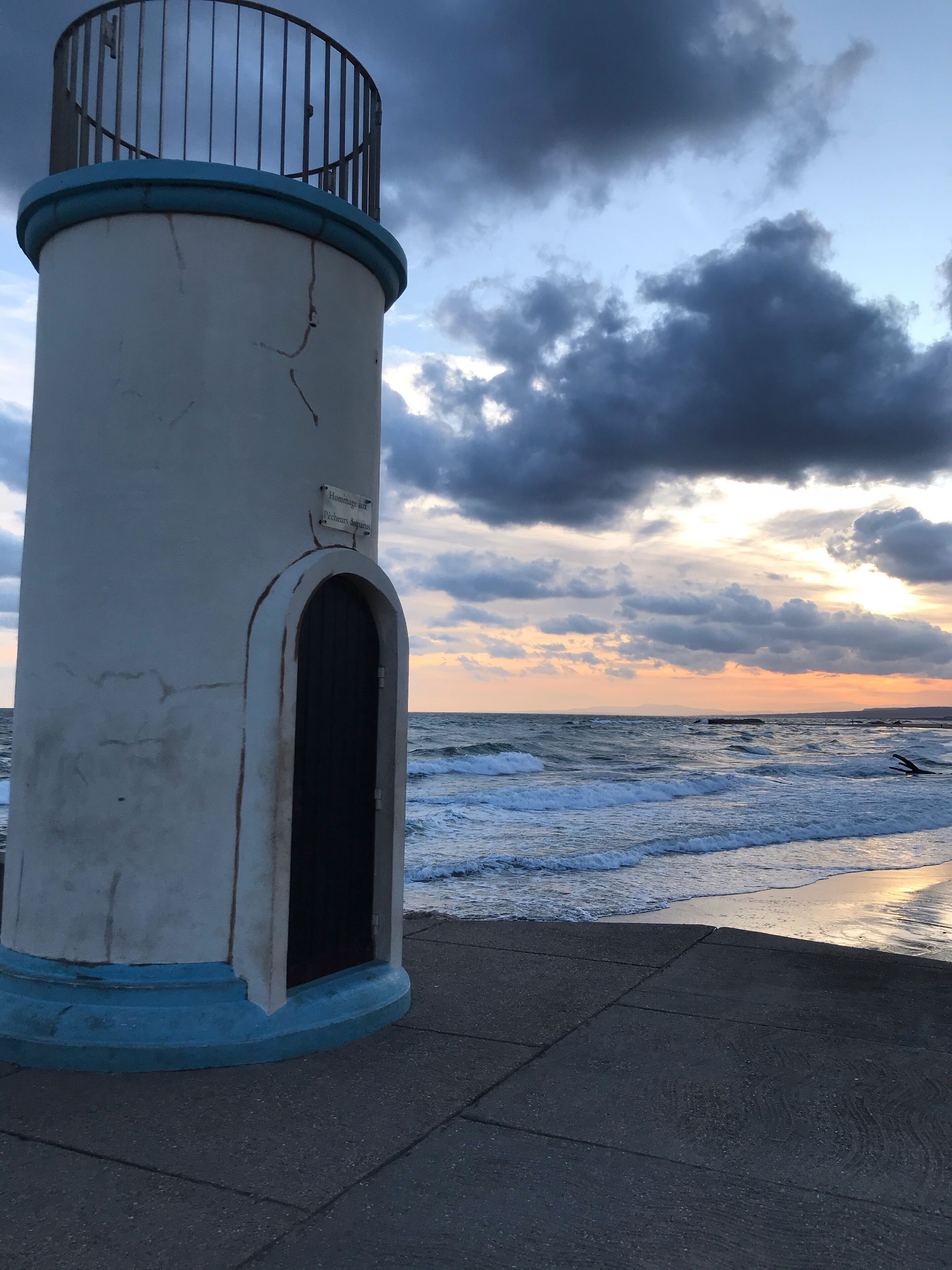 A lighthouse with a metal railing on top, situated on a paved area near the ocean.