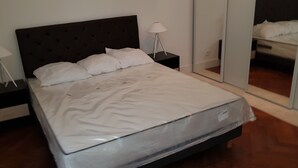 Una cama bien hecha con sábanas blancas en una habitación con suelo de madera. Hay dos mesitas de noche con lámparas y un espejo grande en la pared.