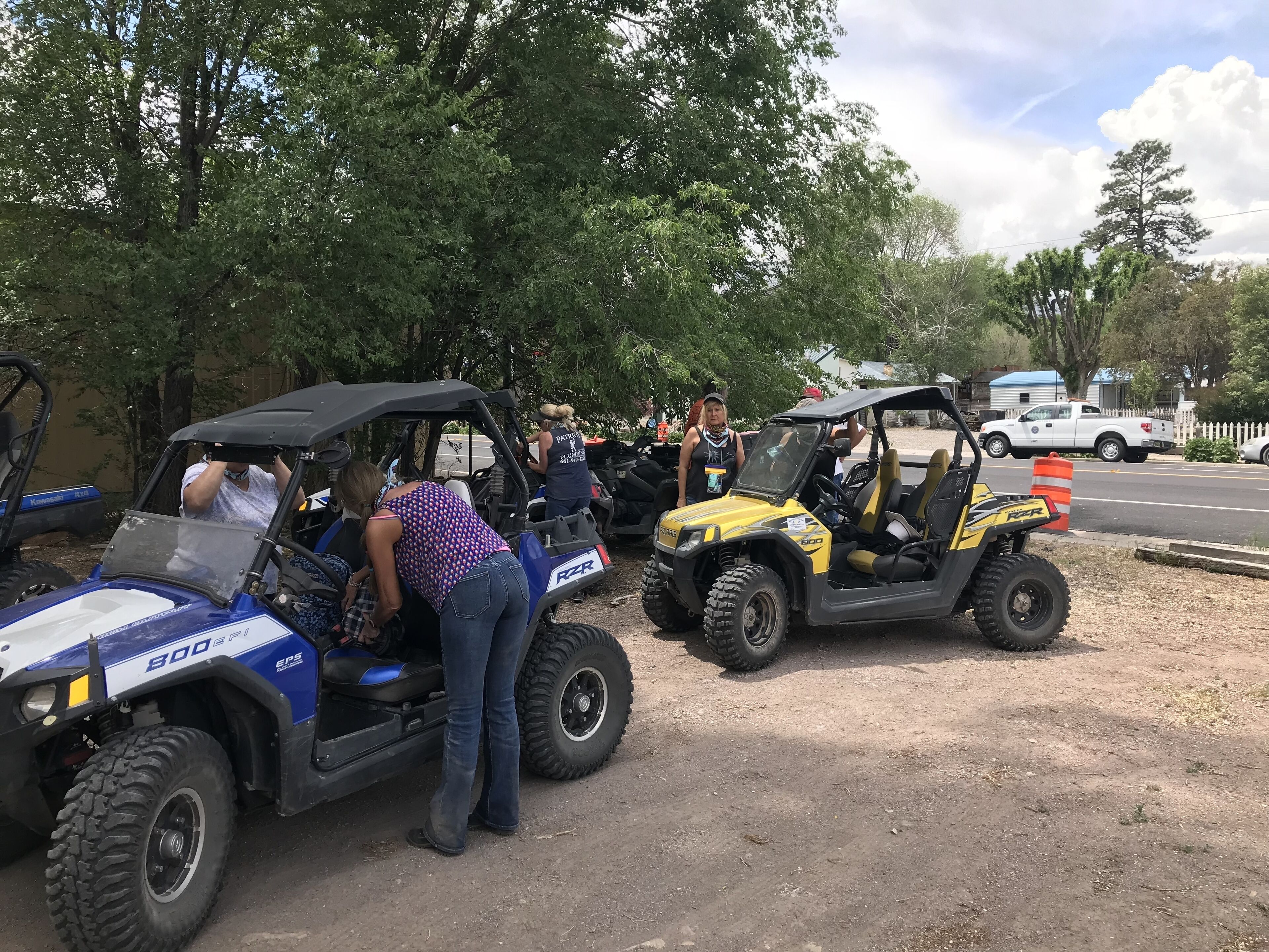 A group of people around off-road vehicles, including a blue and a yellow one, in a parking area with trees and a white pick-up truck in the background.