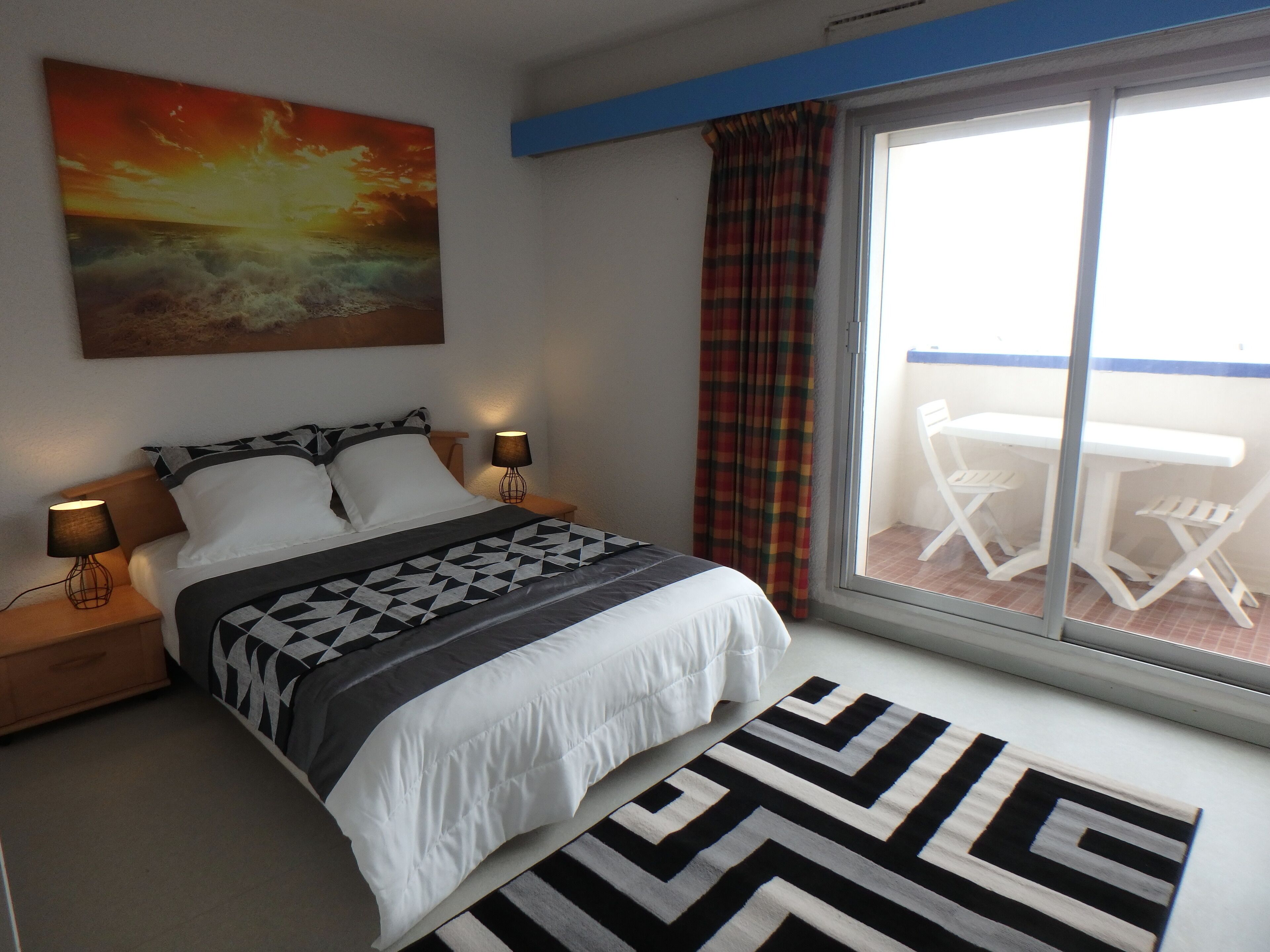 Een slaapkamer met een bed, twee bedlampen, een schilderij, een raam met gordijnen en uitzicht op een buitenruimte met een tafel en stoelen.