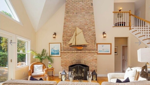A spacious living room with a brick fireplace, a high ceiling, and a staircase leading to an upper level. - Light and Bright East Hampton Home, Perfect Location! (East Hampton)